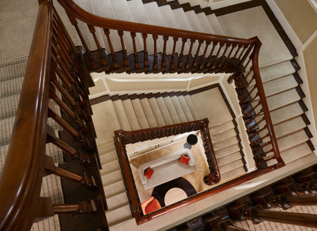 View from above of a grand wooden staircase spiraling downwards, highlighting classic architectural detailing and a cozy lounge area below at master Cannon London apartments.