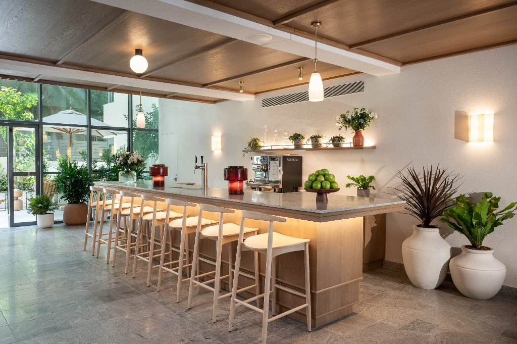 Inviting lounge and coffee bar at master Mazeh with a sleek stone countertop, wooden stools, espresso machine, and abundant greenery in a serene, natural-lit space.